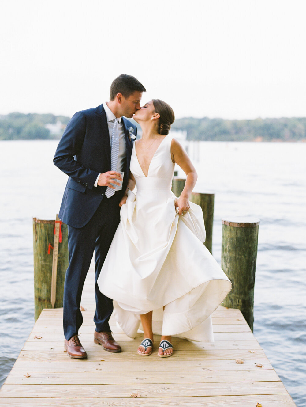 A newlywed couple stands on a wooden dock by the water. The groom, in a dark suit, holds a drink and kisses the bride, who wears a white dress and holds its hem slightly lifted to reveal casual sandals. The background shows a calm lake with a forested shoreline.