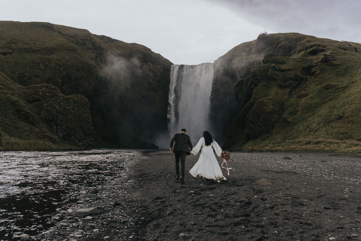 Husband and wife walk hand in hand toward an Iceland waterfall