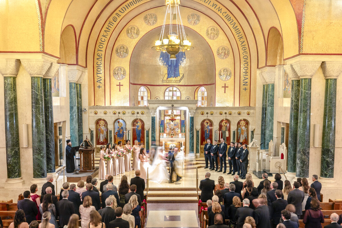 A wedding ceremony inside a large, ornate church. The bride and groom stand surrounded by the wedding party. Guests are seated in pews, and the church features arches, pillars, religious icons, and a grand chandelier.