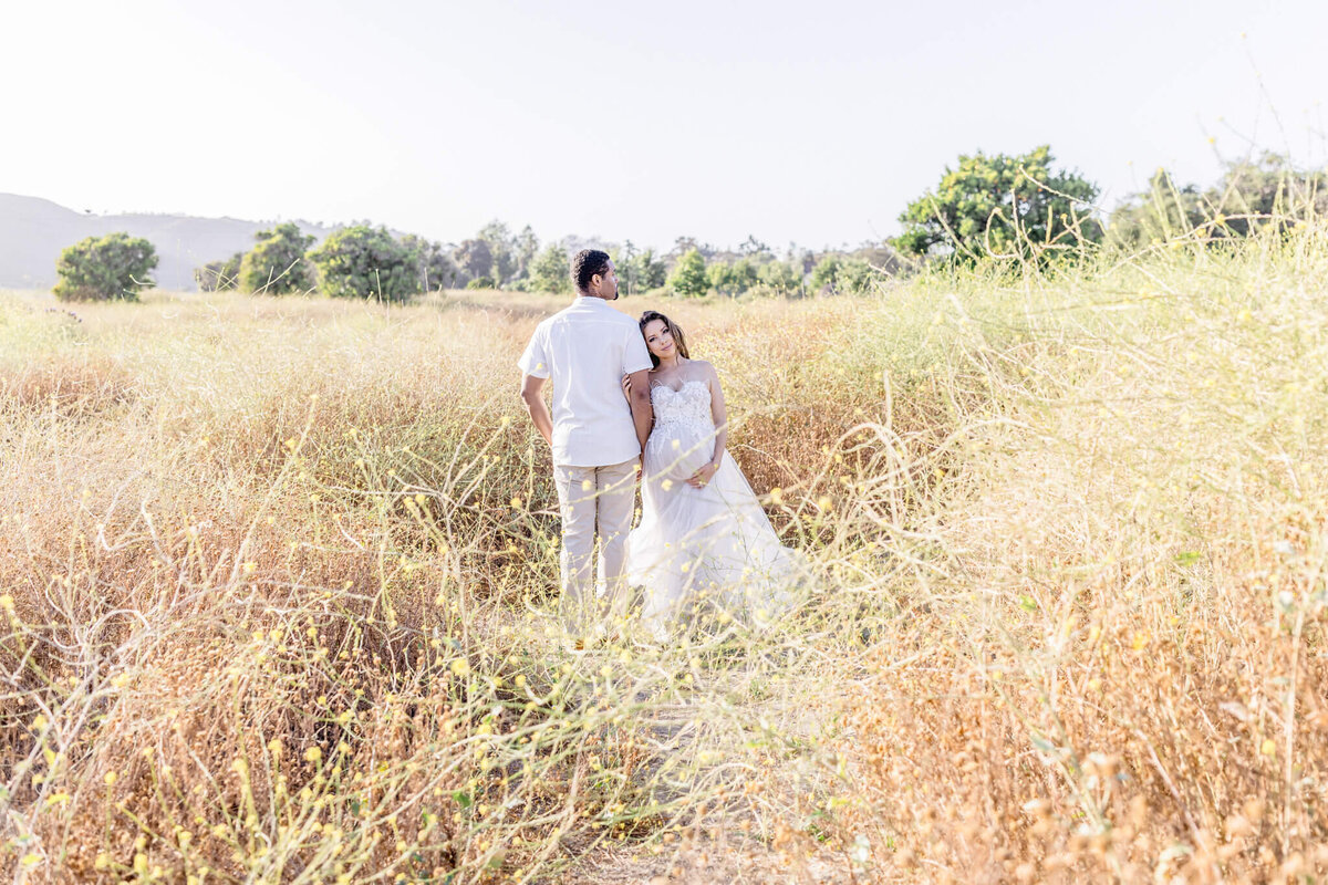 A couple stands in a sunlit field of tall, dry grass. The woman, in a white dress, leans affectionately on the man, who wears a white shirt and trousers. Trees and hills are visible in the distance under a clear sky.