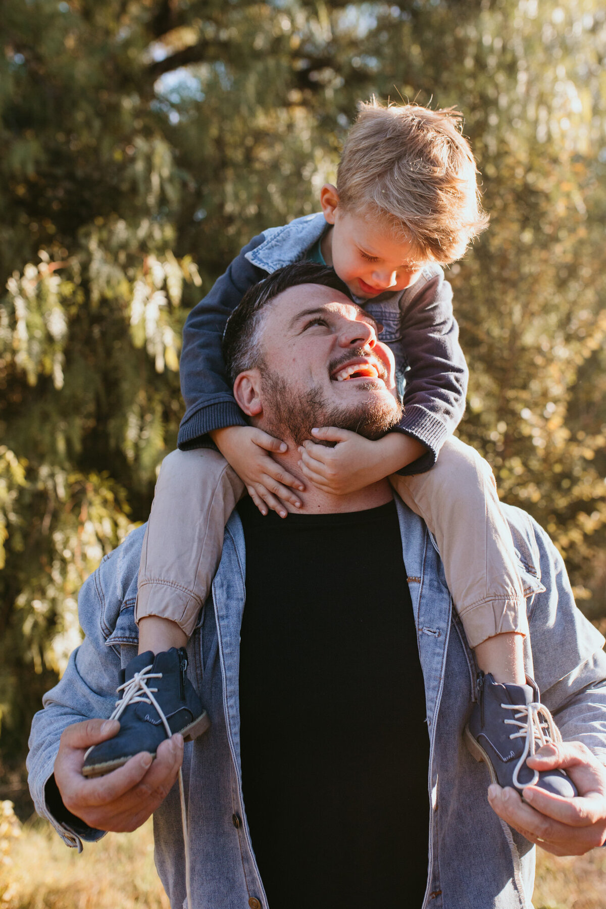 A blond boy is sitting on his Dad's shoulders while bending down to kiss him. 