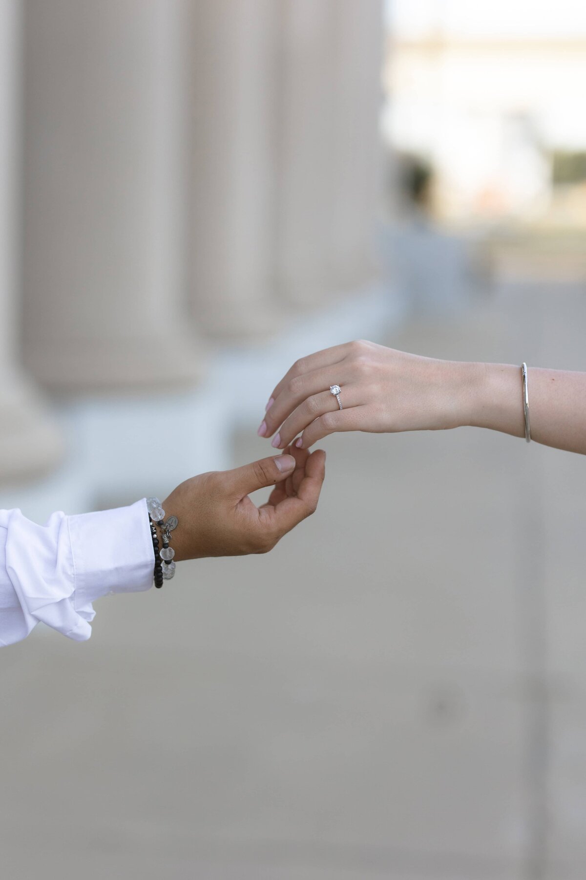 A detailed close-up of an engaged couple gently reaching for each other’s hands, highlighting the bride-to-be’s delicate diamond ring and simple bracelet. The background features soft architectural columns and a shallow depth of field, creating a romantic and refined aesthetic. Captured by Heartful Films, a Jacksonville engagement photographer and videographer known for capturing elegant, emotion-filled details that tell a love story.