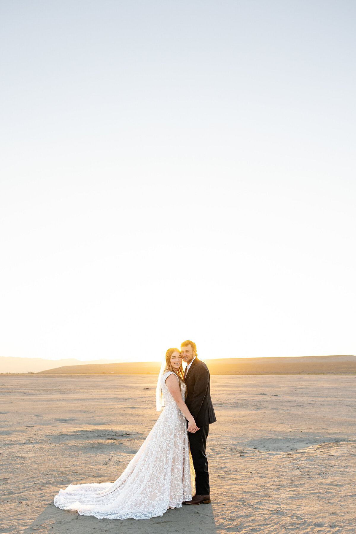 Bride and groom smile at the camera, standing together with the sun rising over the Salt Flats.