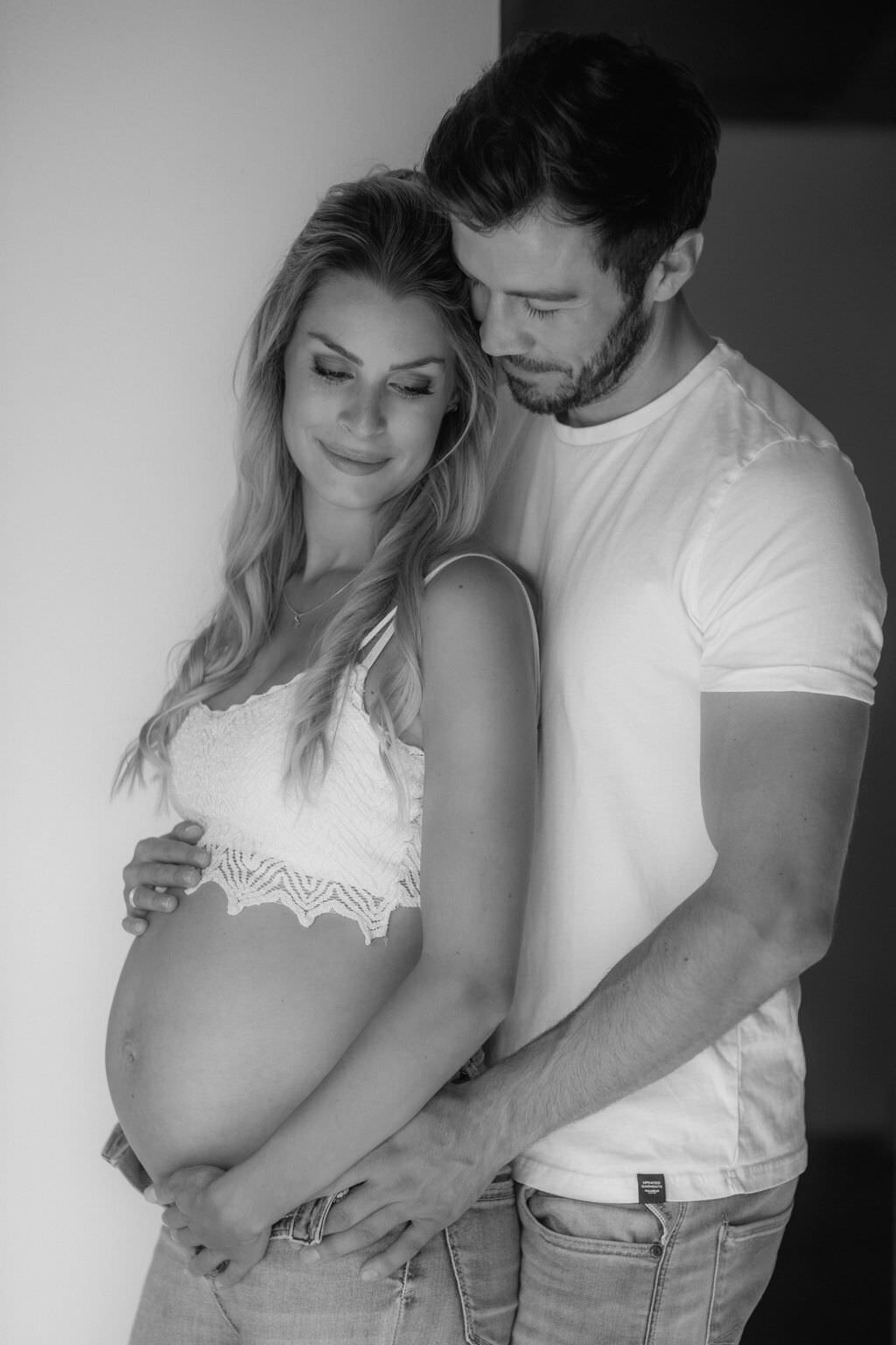 An expecting couple stands in a warm embrace holding the baby bump.