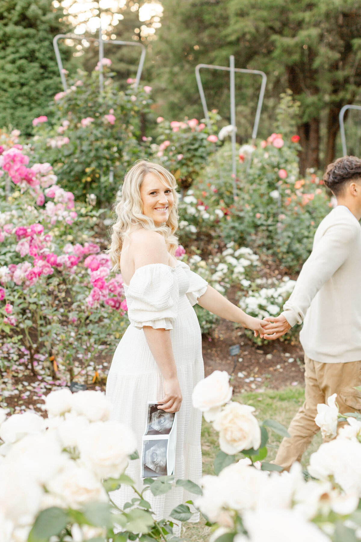 Woman and Man in neutral clothes hold hands while walking through the Portland Rose Gardens. Woman is looking at the camera and holding ultrasound photos down by her side.