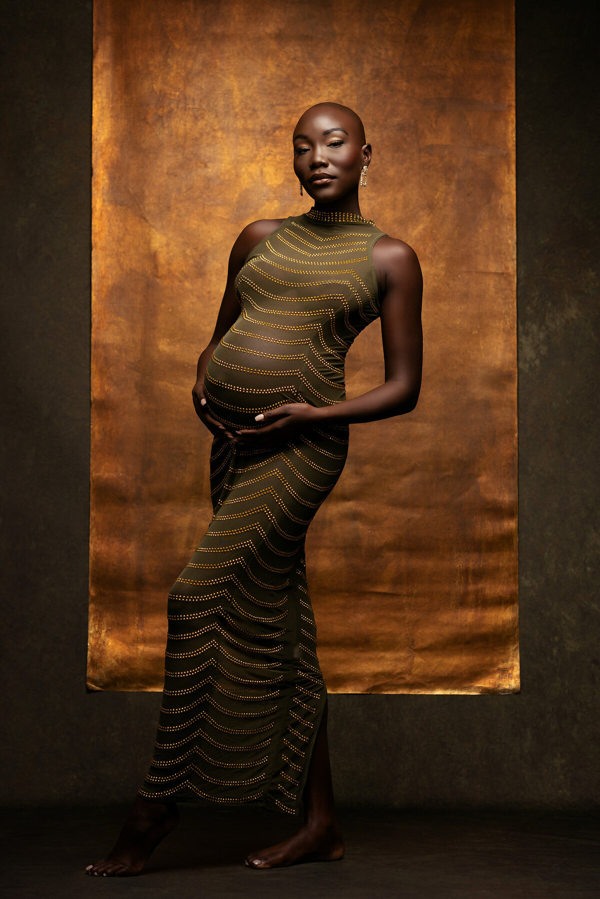 A confident woman stands elegantly before a rich golden backdrop, wearing a stunning olive-green dress adorned with delicate golden accents that gracefully outline her figure, captured by Dallas maternity photographer Lindsay Walden.