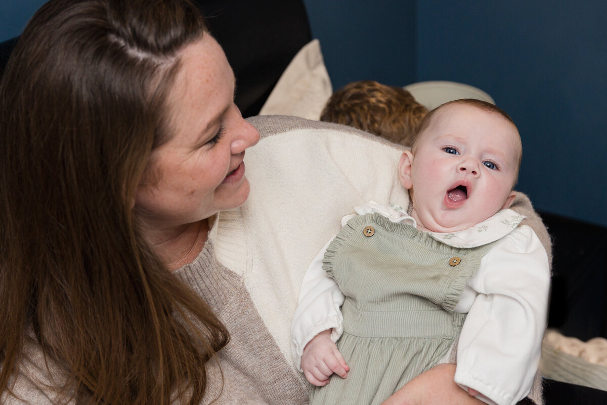 A woman smiling at a baby in her arms who is wearing a green outfit and looking amused.