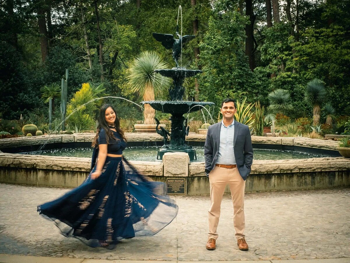 A woman in a flowing blue dress twirling in front of a fountain with a man smiling