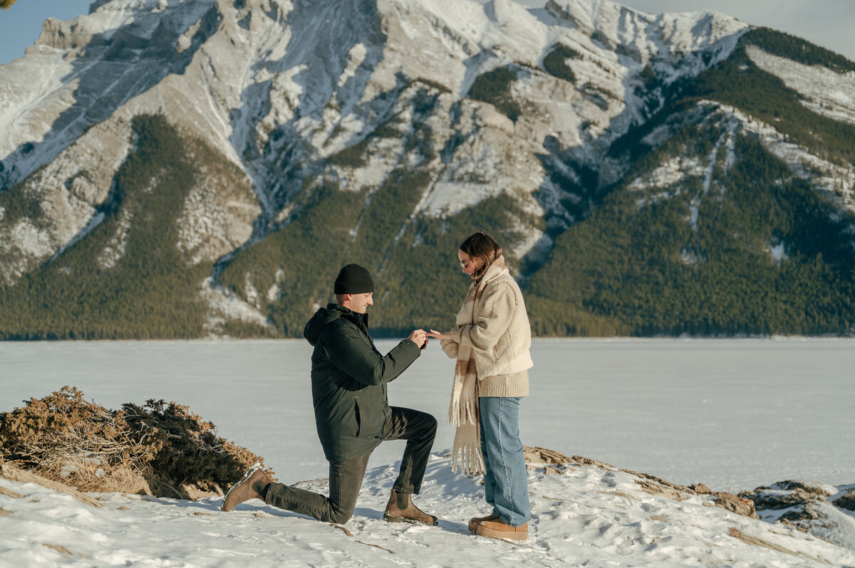 Surprise proposal at Lake Minnewanka as man kneels in front of mountain views with snow-dusted trees and sunshine lighting the moment.