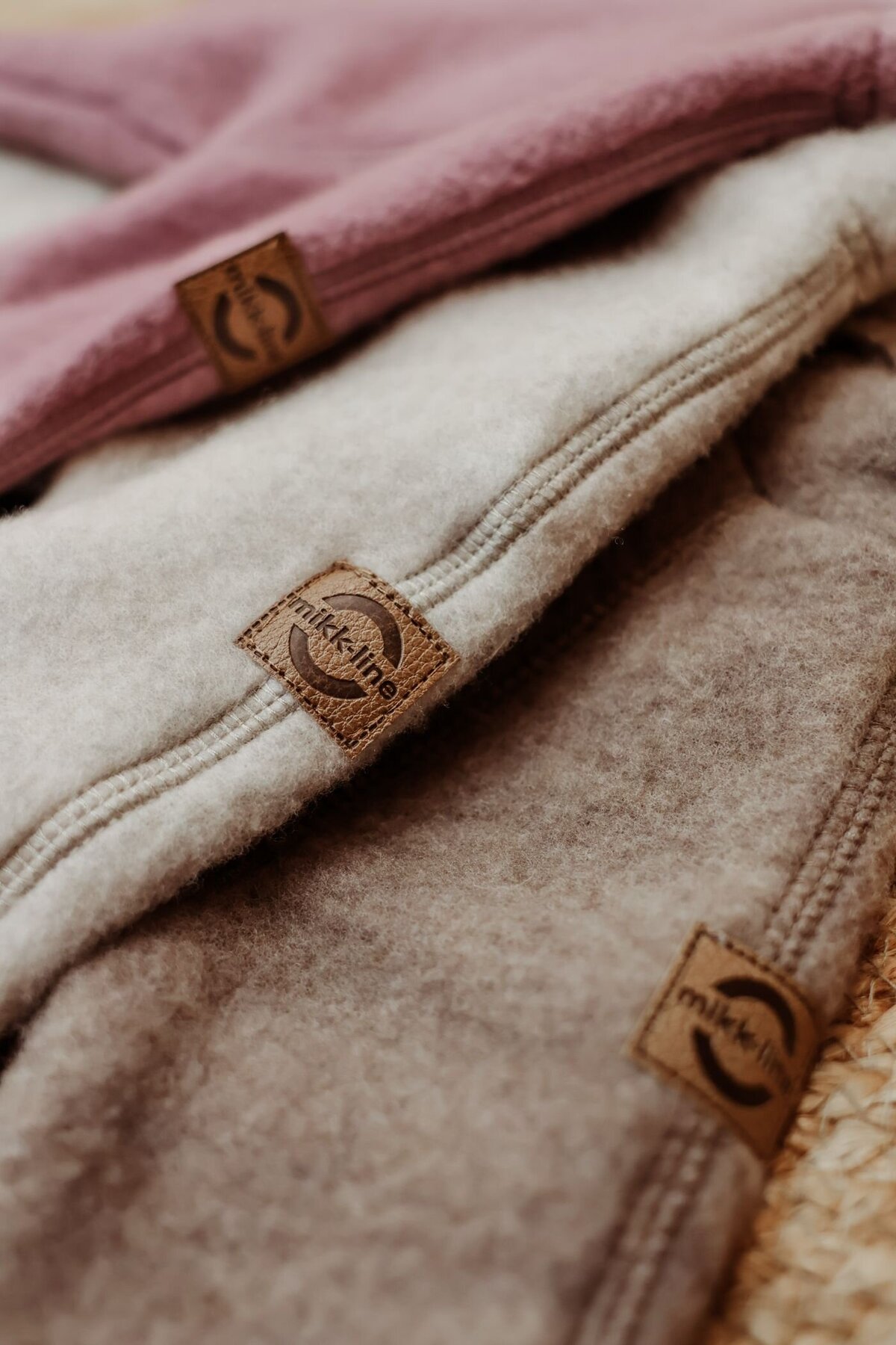 Drie opgevouwen stukken zachte, wollen stof in verschillende kleuren (roze, lichtgrijs en bruin) met een bruin leren label met het opschrift "mikk-line" in het midden