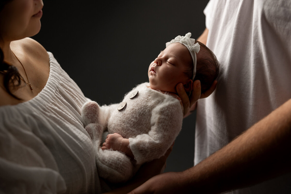 Newborn baby girl peacefully resting in her parent's arms, wearing a soft white knit outfit with button details and a sweet headband, against a dark backdrop. Captured by Inland Empire newborn photographer Kaitlyn Dawn Photography.