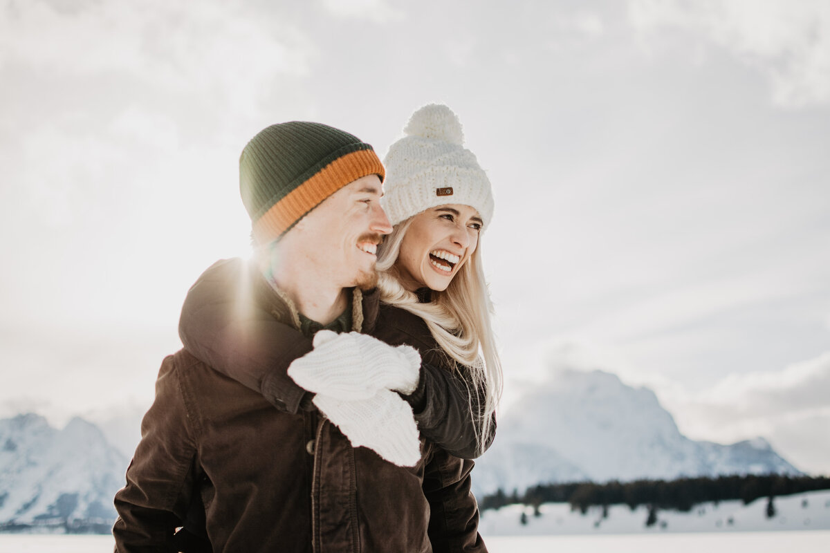 jackson hole photographers captures man and woman laughing as the sun beams behind them for their winter engagement session