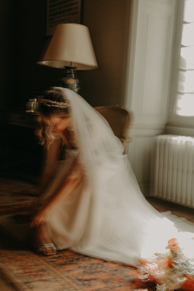 Mariée prise en mouvement, effet de flou, ajustant sa chaussure dans un décor feutré. Immortalisée par Laura, photographe professionnelle en Vendée.