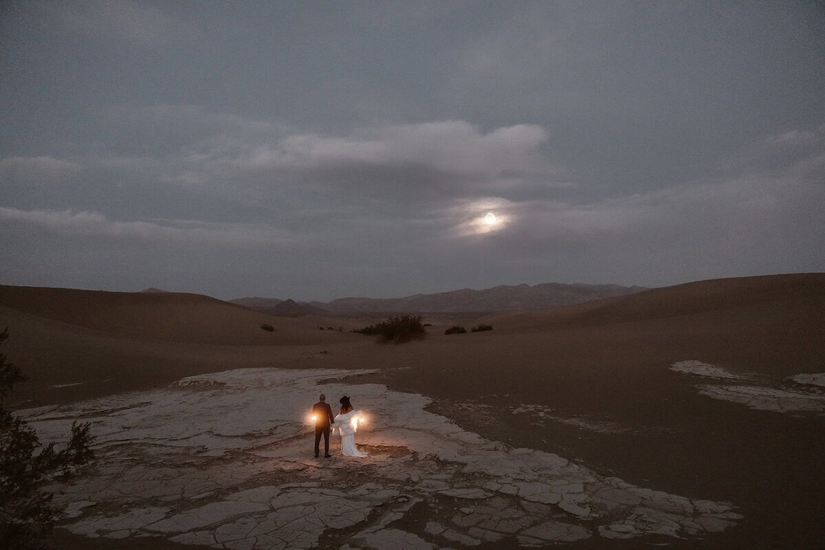 Couple holding lanterns while walking through cracked desert ground under a cloudy night sky with the moon glowing above distant mountains.