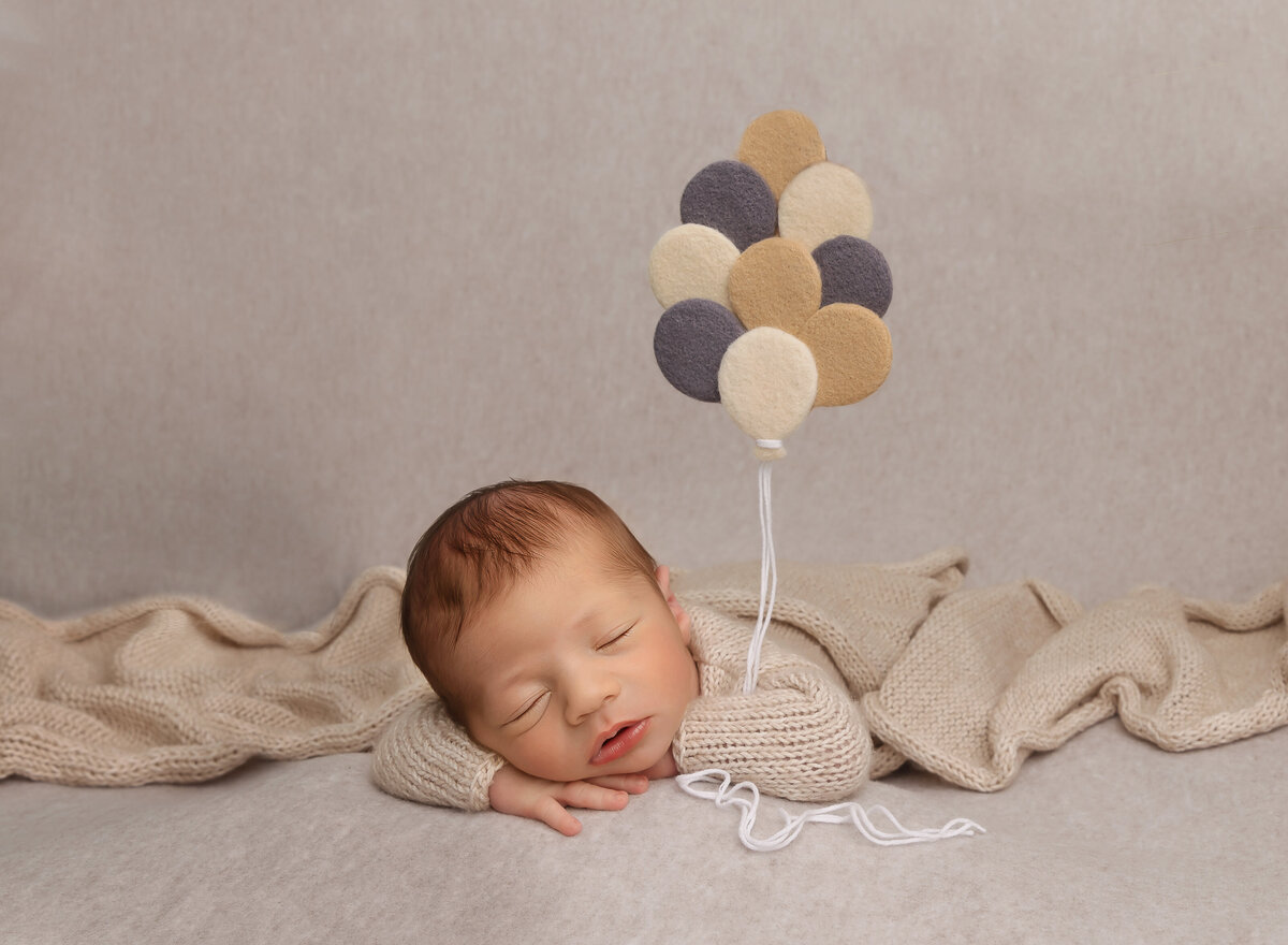 Baby boy in a beige knit onesie is laying on his belly with one hand folded under his chin. the other arm is bent to give the illusion that he is holding suspended felt balloons. Captured by top Brooklyn newborn photographer Chaya Bornstein