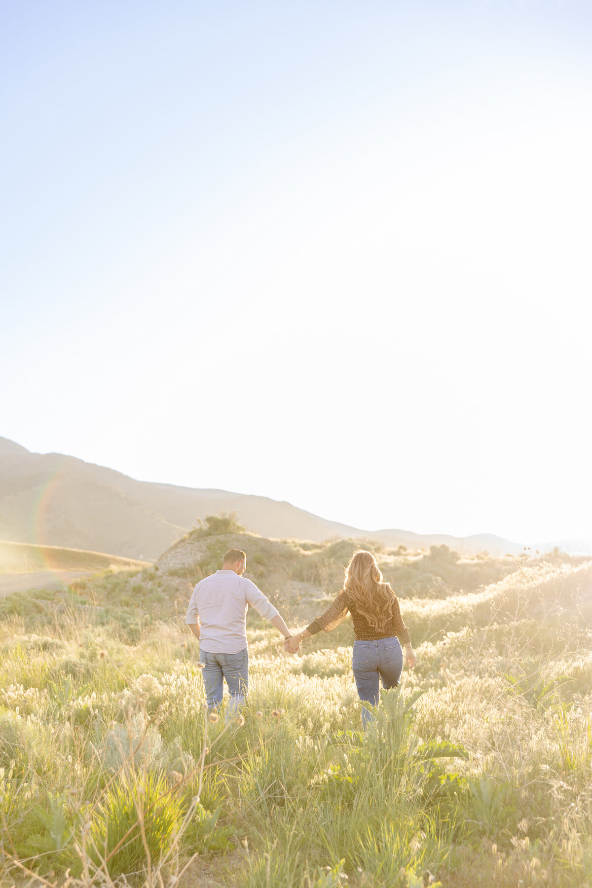 A couple walks hand in hand through a sunlit field in Saratoga Springs, Utah. The warm golden hour light creates a dreamy atmosphere as they journey together toward the horizon.