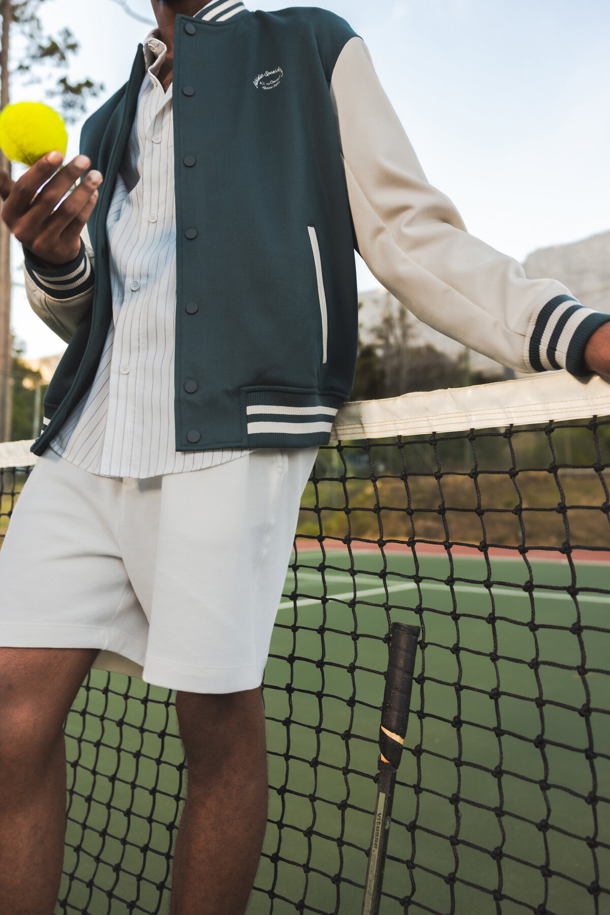 Cropped view of a man in a sporty varsity jacket and white shorts leaning on a tennis net, holding a yellow tennis ball on an outdoor court.