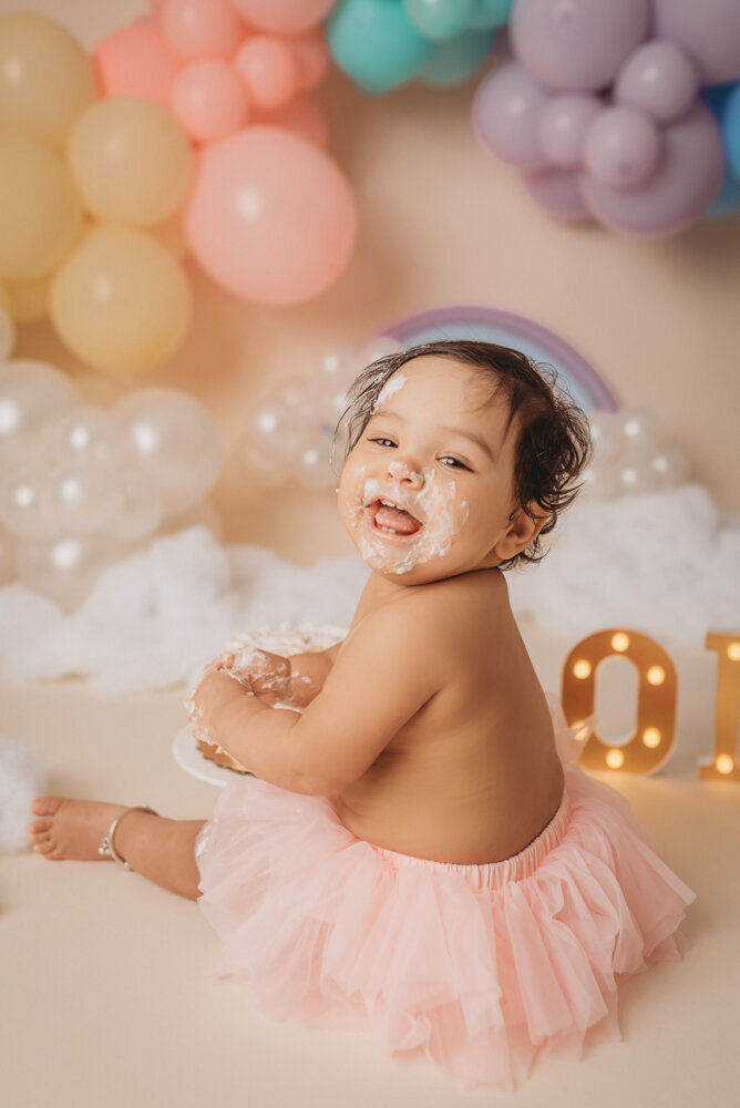Photo shoot at Atlanta, Georgia cake smash photographer studio with one year old little girl in pink tutu smashing a cake with rainbow pastel balloon garland behind her with white fluffy clouds.