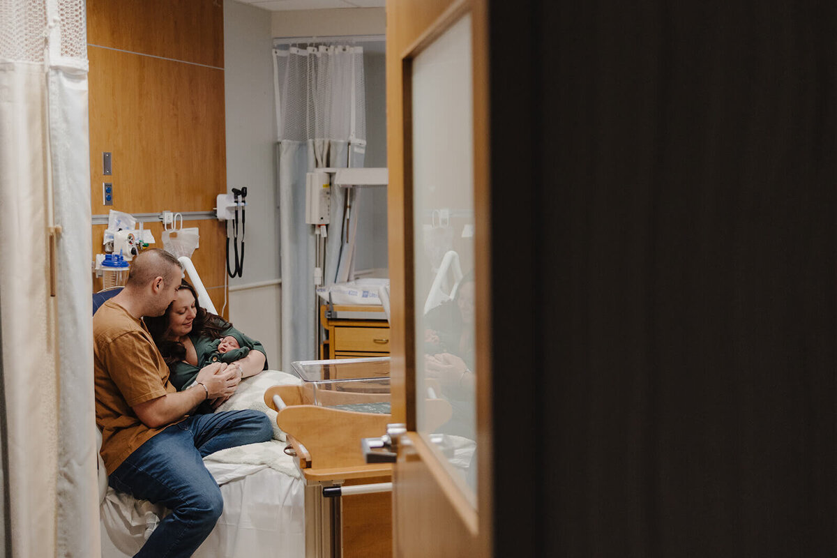 Couple looking at baby as they sit on a bead at  Finger Lakes Maternity session