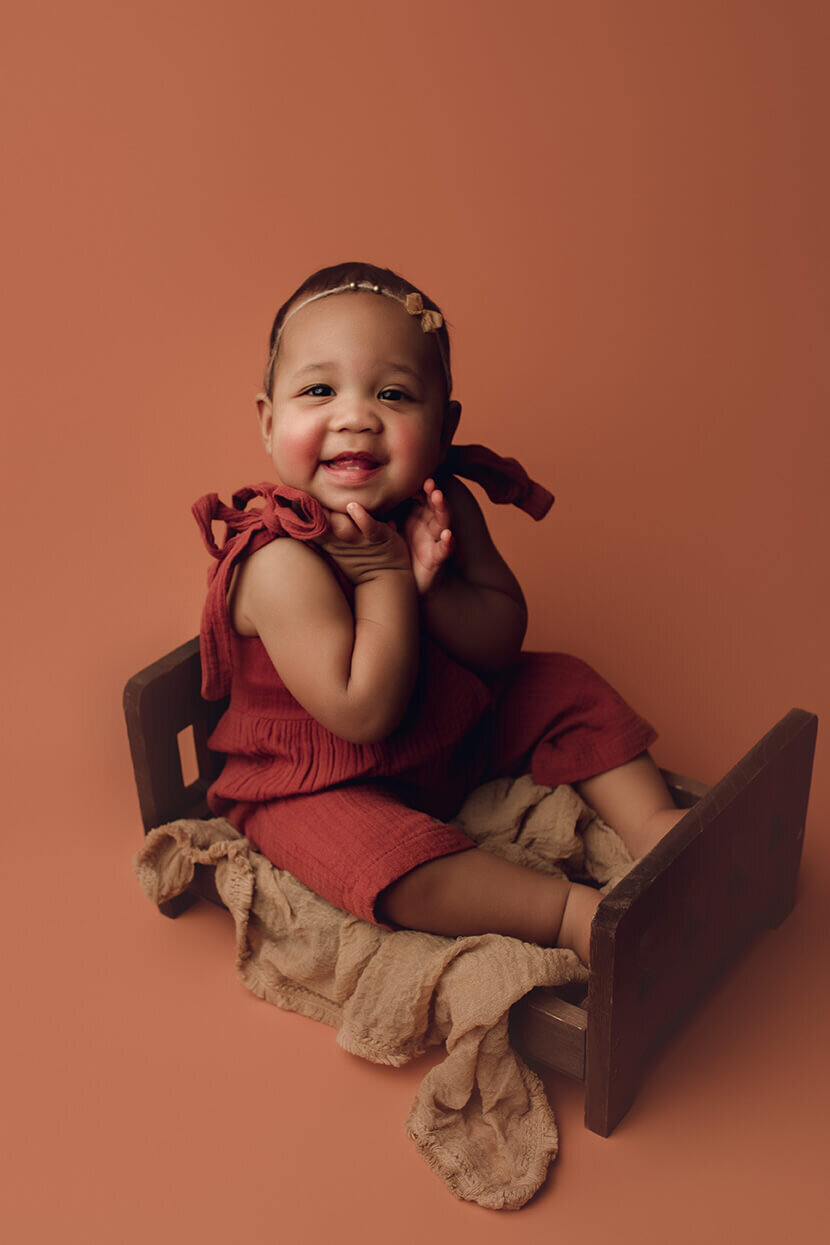 a one year old girl holding her face and smiling on a wooden bed