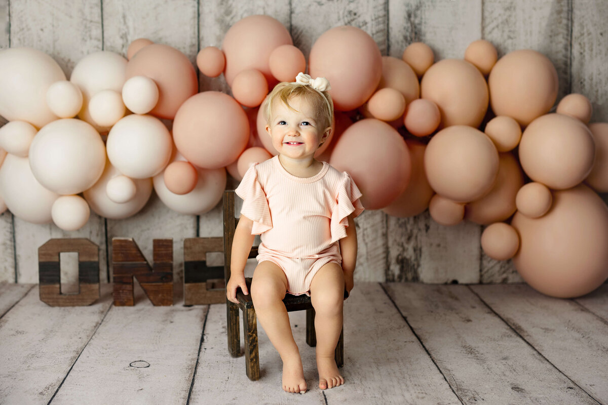 one year old girl sitting on a white wooden chair with wooden one letter and pink balloons behind her during her one year old session