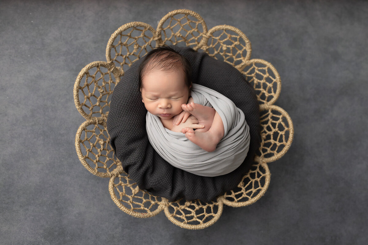 Newborn baby swaddled in soft gray fabric, lying peacefully with toes peeking out on a textured dark cushion and decorative flower-shaped woven prop, styled by a Dallas newborn photographer.