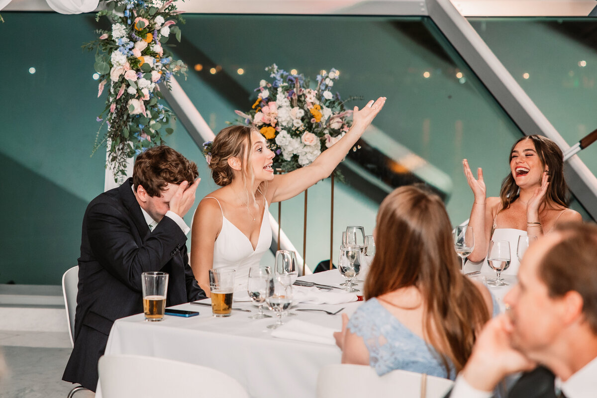A groom puts a palm on his face as his bride makes wild gestures at head table