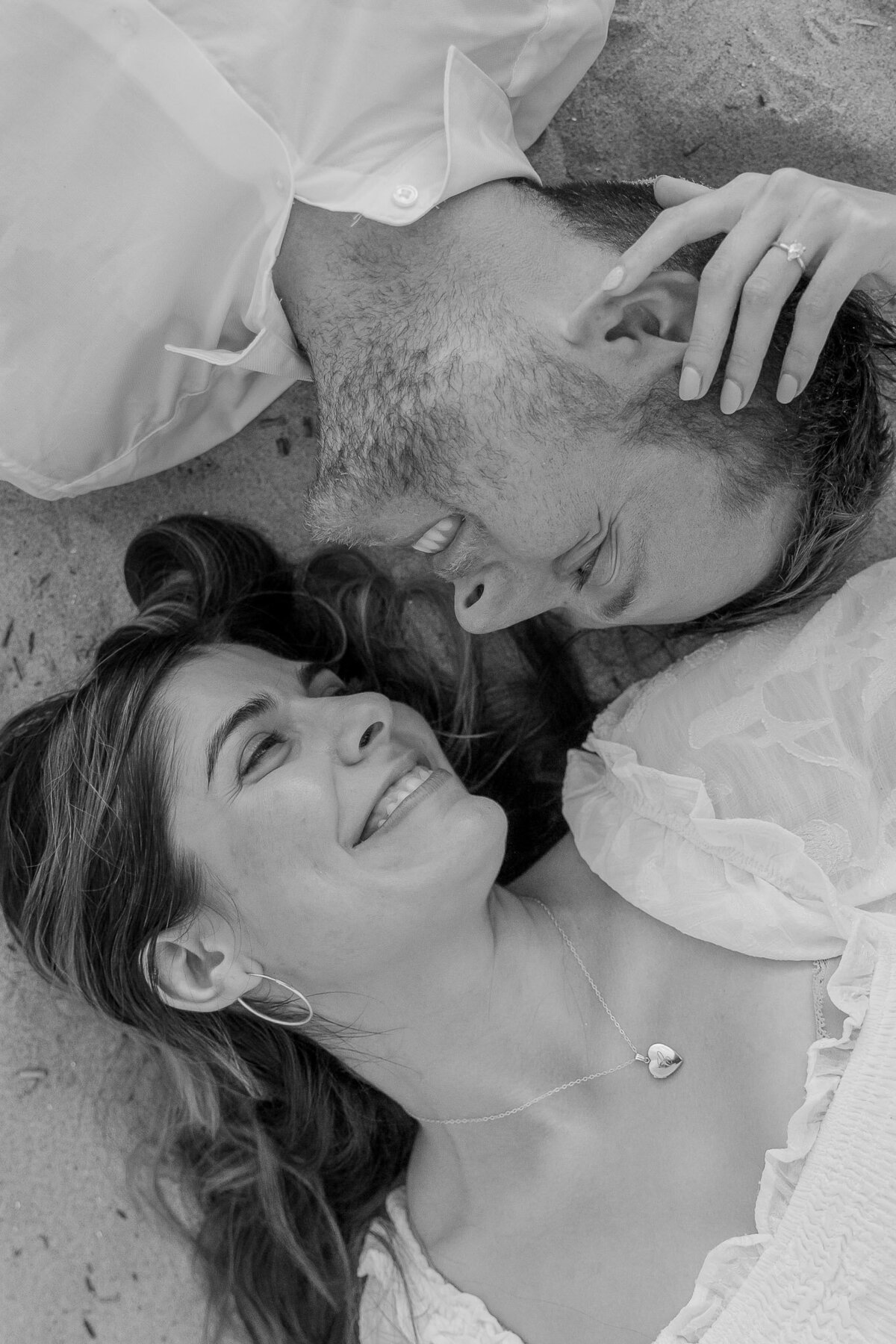 A bird's eye view of a couple laying on the ground opposite of each other. The newly engaged couple are looking into each other's eyes and smiling big.