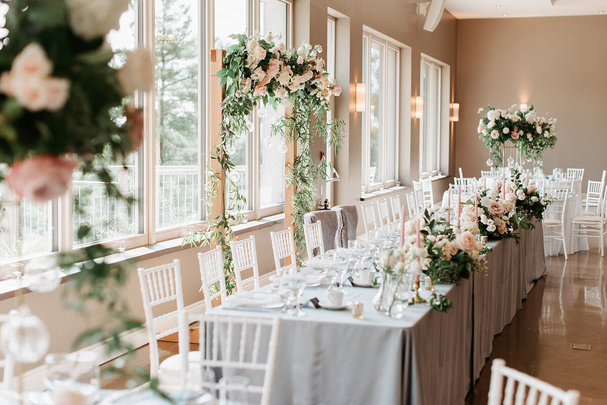 In front of a large glass window, a long head table with dusty blue linens and blush centrepieces sits in front of a large wood arch covered in romantic flowers set for a wedding at Le Belvedere venue in Wakefield Quebec