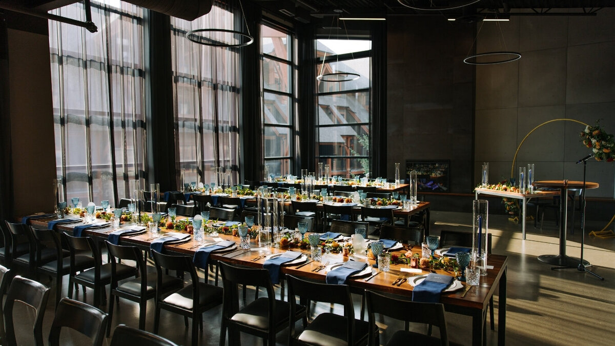 Interior of Mainspace Canmore set for a summer wedding reception featuring the Dark Industrial décor package — long wooden tables with navy napkins, blue glassware, and candlelit centerpieces under modern ring lighting and floor-to-ceiling windows with mountain views.