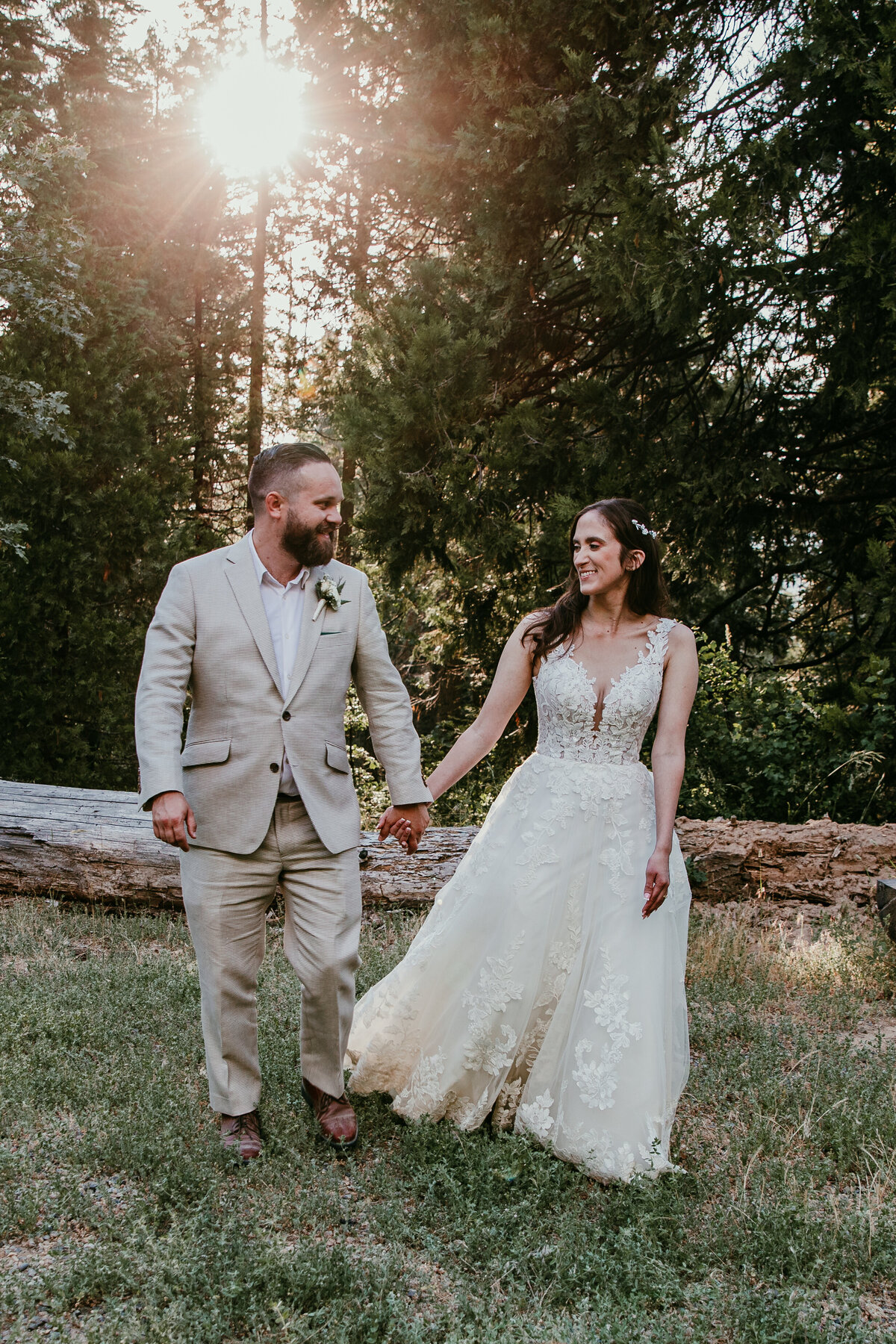 Bride and groom walking hand in hand through a sunlit forest near Yosemite, with golden light filtering through the trees.