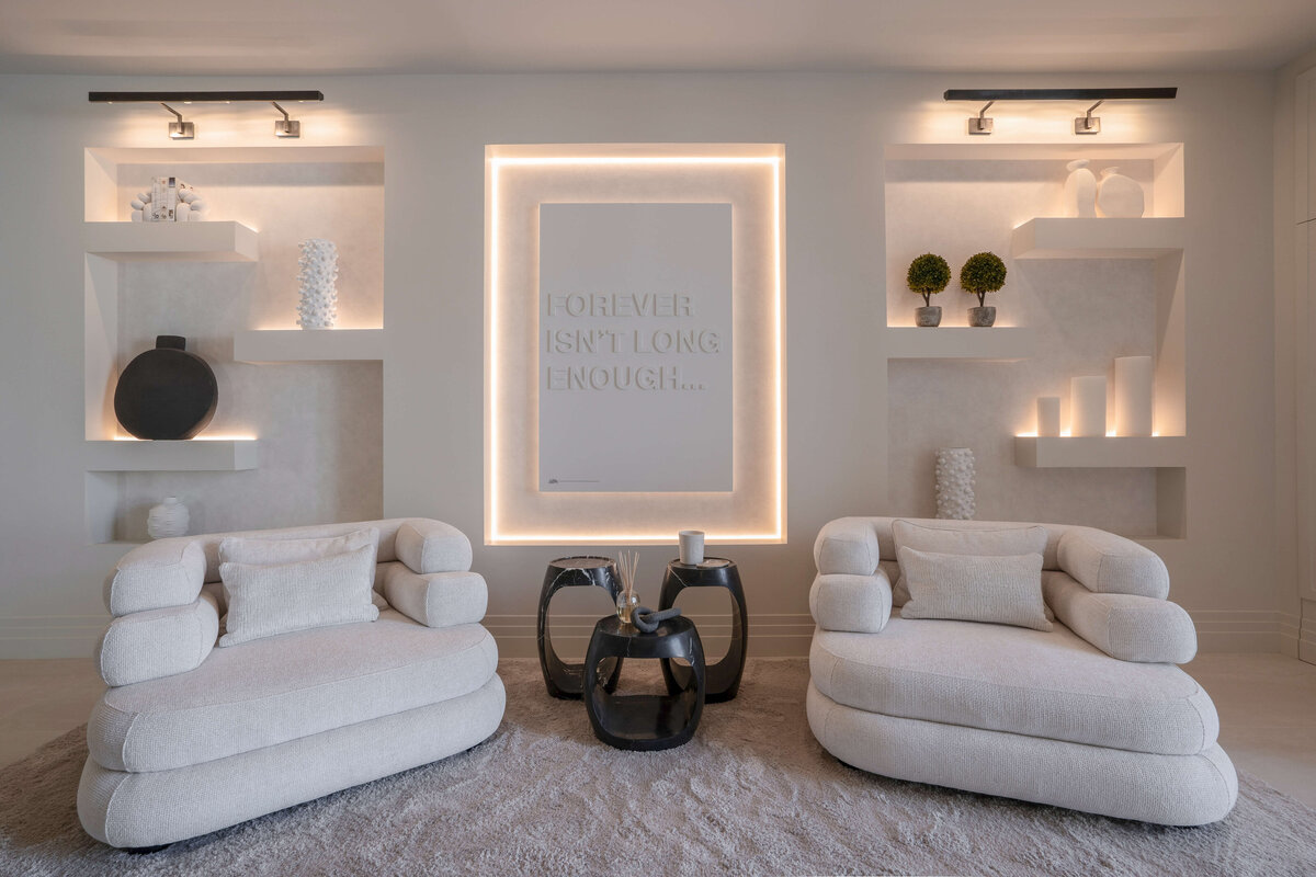 A spacious, modern entry area at the foot of the stairs, with a focus on light, neutral tones. There are two plush, light-colored armchairs and a soft, textured rug. Built-in shelves with decorative items and a wall with the illuminated text 'FOREVER ISN'T LONG ENOUGH' add a sleek coolness to the space. In the background, a dining area with a large table and pendant lights is visible through glass doors.