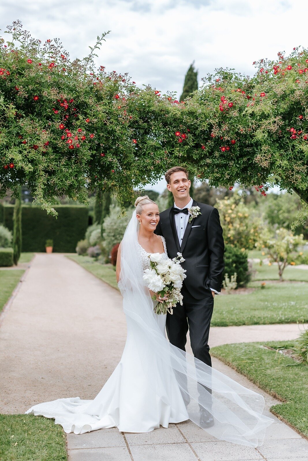 A couple getting married at Coombe Estate in the Yarra Valley, standing in manicured gardens and holding a white bouquet.