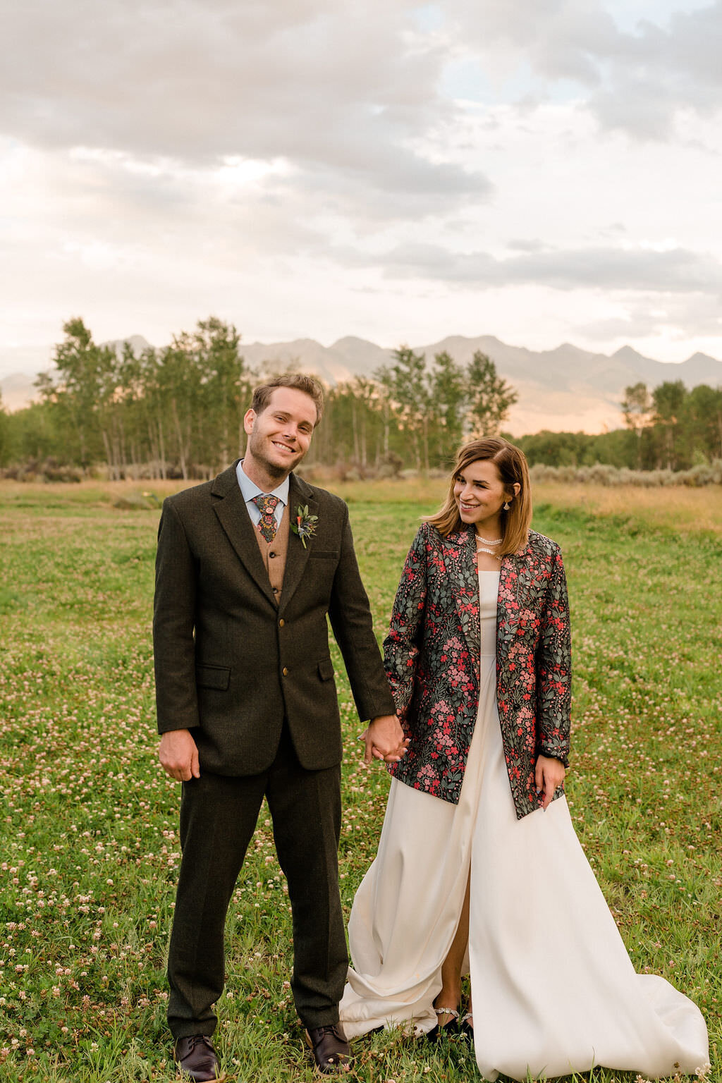 bride with a colorful jacket over her dress holding her grooms hand as they both smile