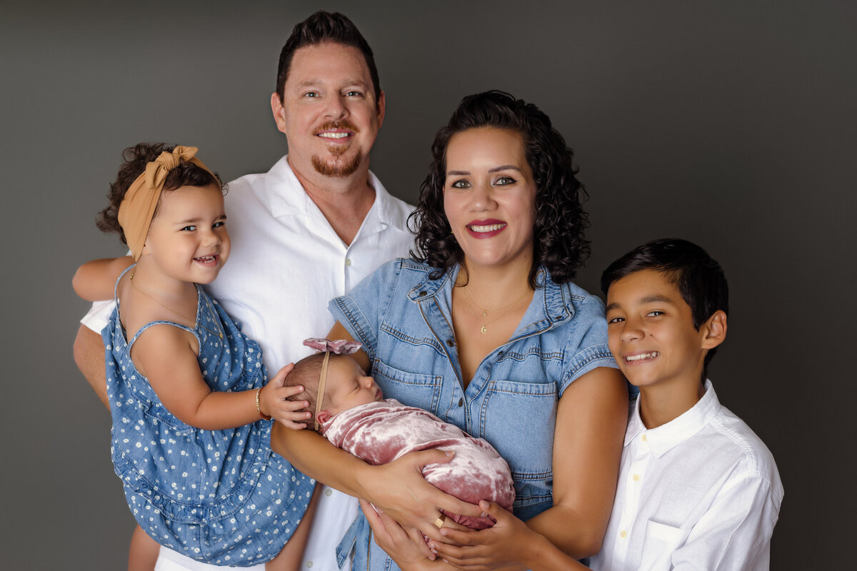 Newborn Photographer, a young family stands together, mom holds baby and two siblings stand nearby