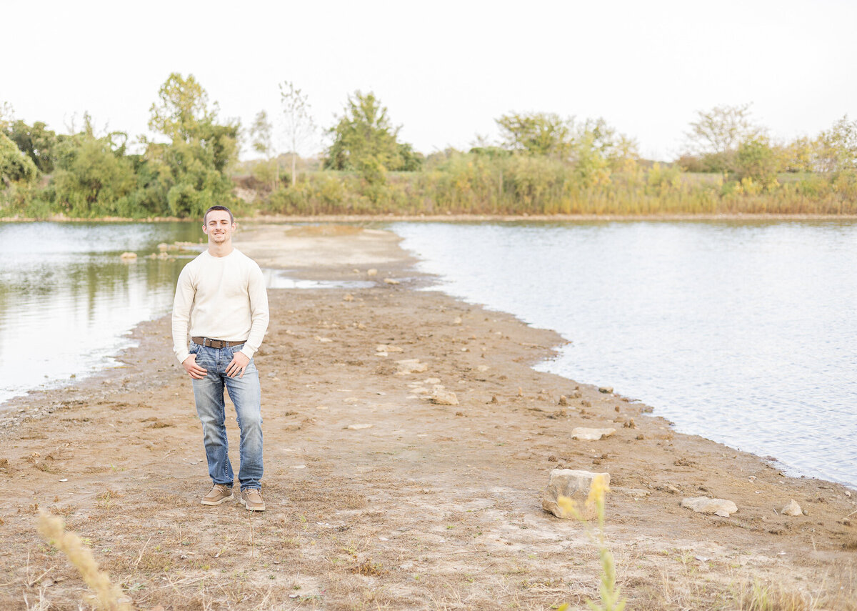 high school senior male, standing on dry land in the middle of a pond, with trees in the background, thumbs in his pockets, wearing a light tan long sleeve shirt, blue jeans, a belt, and tan dress shoes, smiling at the camera