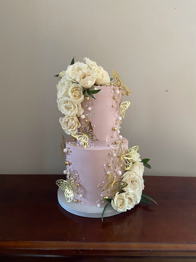 Photo shows a small two tier cutting cake with pastel purple frosting. On the top left and bottom right are small cascades of white roses. Lining the inside of each cluster are white and gold sugar pearls, with gold butterflies as accents.
