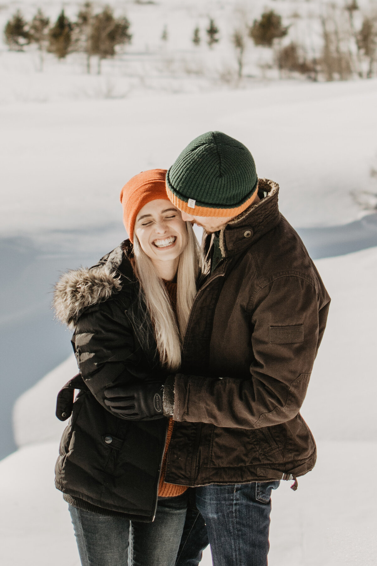 winter engagement photos with man holding woman in a snowy field in Jackson Hole as she laughs during their Grand Teton  winter engagement session captured by jackson hole photographers