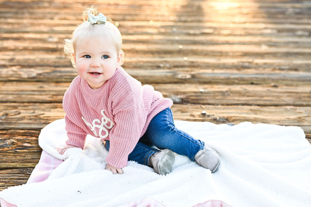A baby smiling and sitting on a blanket on a wooden deck.