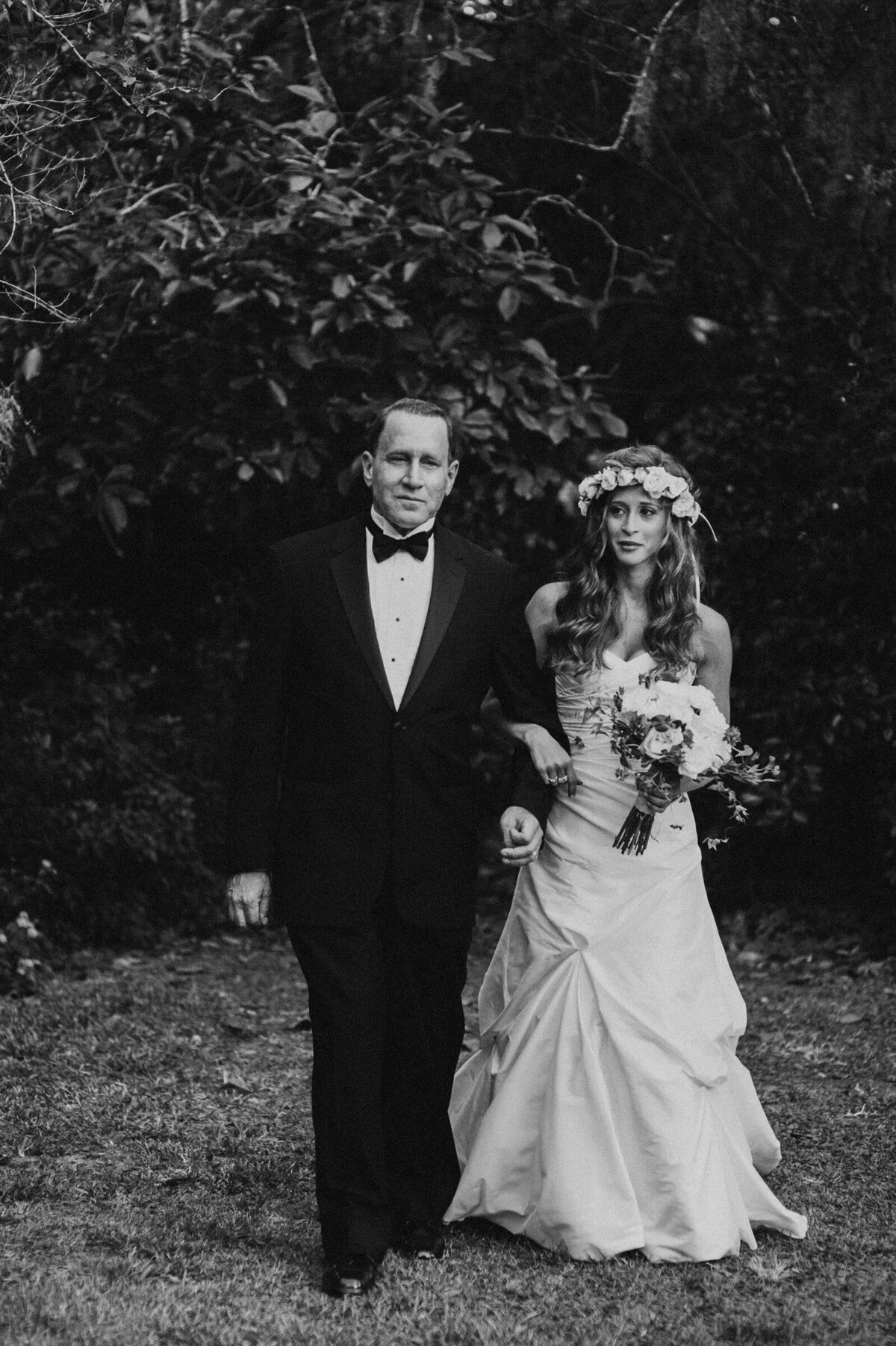 A black and white photo of a bride walking arm in arm with an older man through a garden