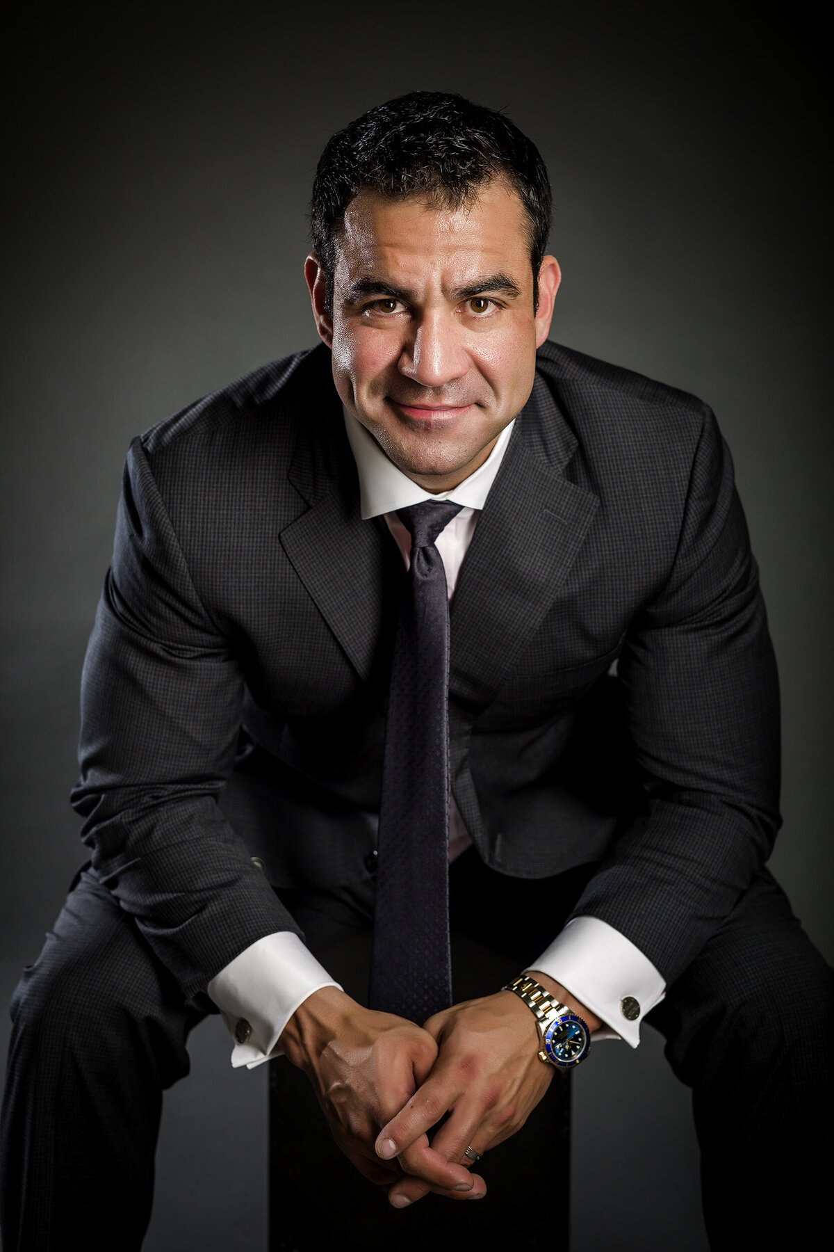 Male headshot client wearing a suit, sitting & leaning forward with hands clasped in his lap & smiling at the camera