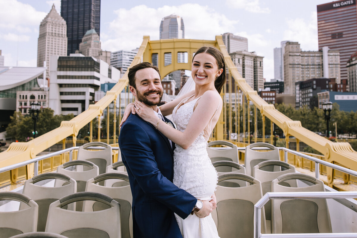 Bride and groom cheering on a bus rooftop driving over the Andy Warhol Bridge with the Pittsburgh skyline behind them. 