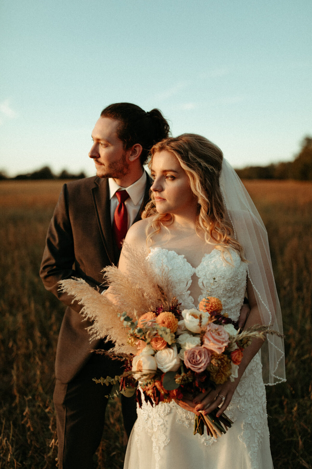 Bride in wedding dress and veil holding floral bouquet with pampas grass standing in front of the groom in grey suit in a field watching sunset