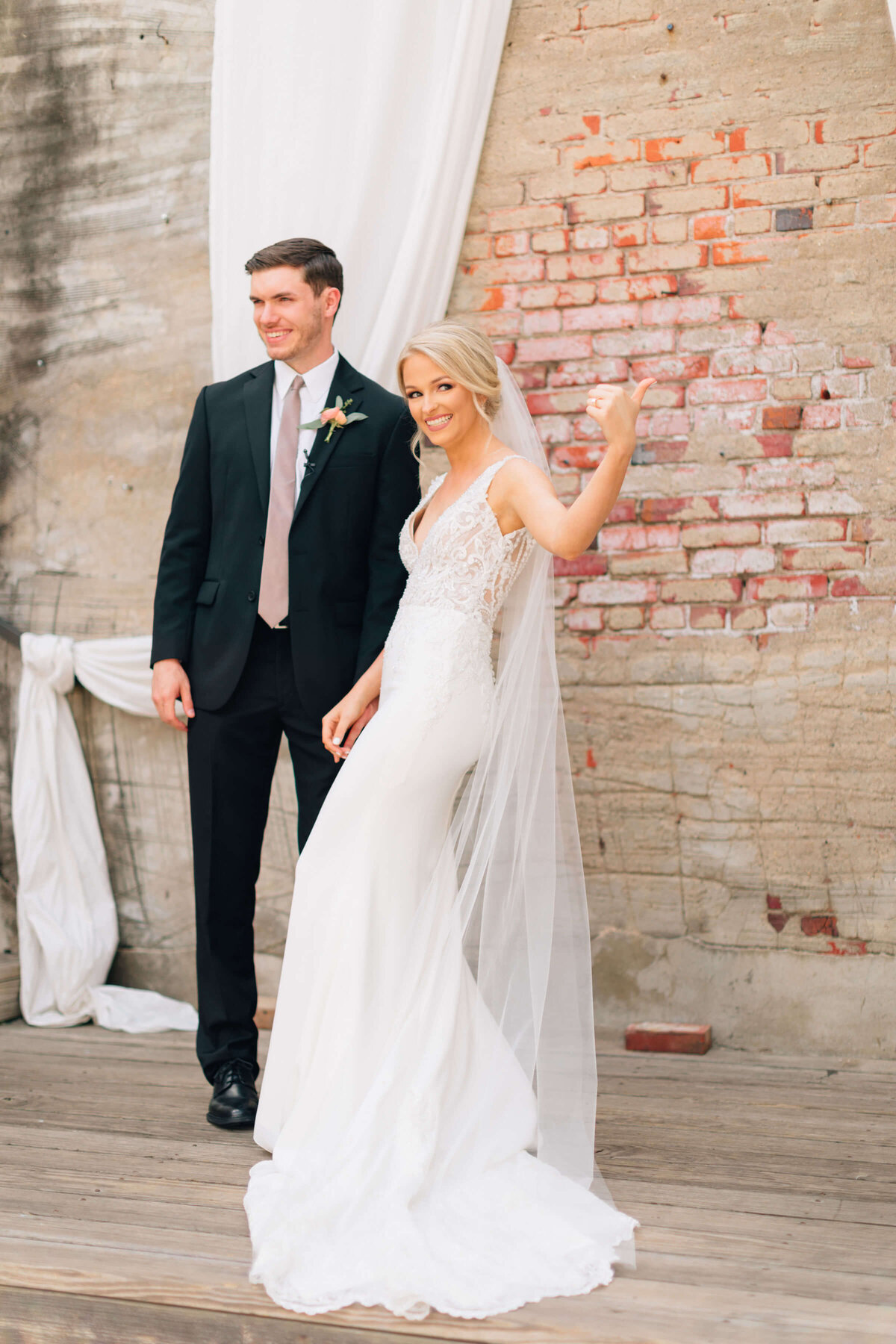 A bride in a white dress and veil smiles and gives a thumbs up next to a groom in a dark suit, standing on wooden floorboards in front of a brick wall with white drapery.