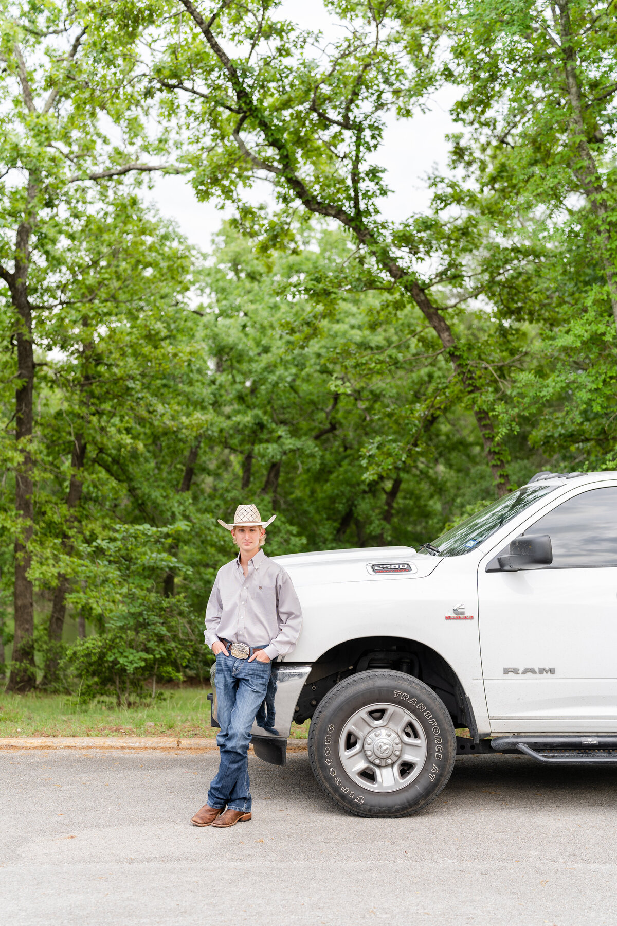 High School senior guy leaning on front of truck smiling at camera and hands in pocket while wearing button down, cowboy hat and jeans