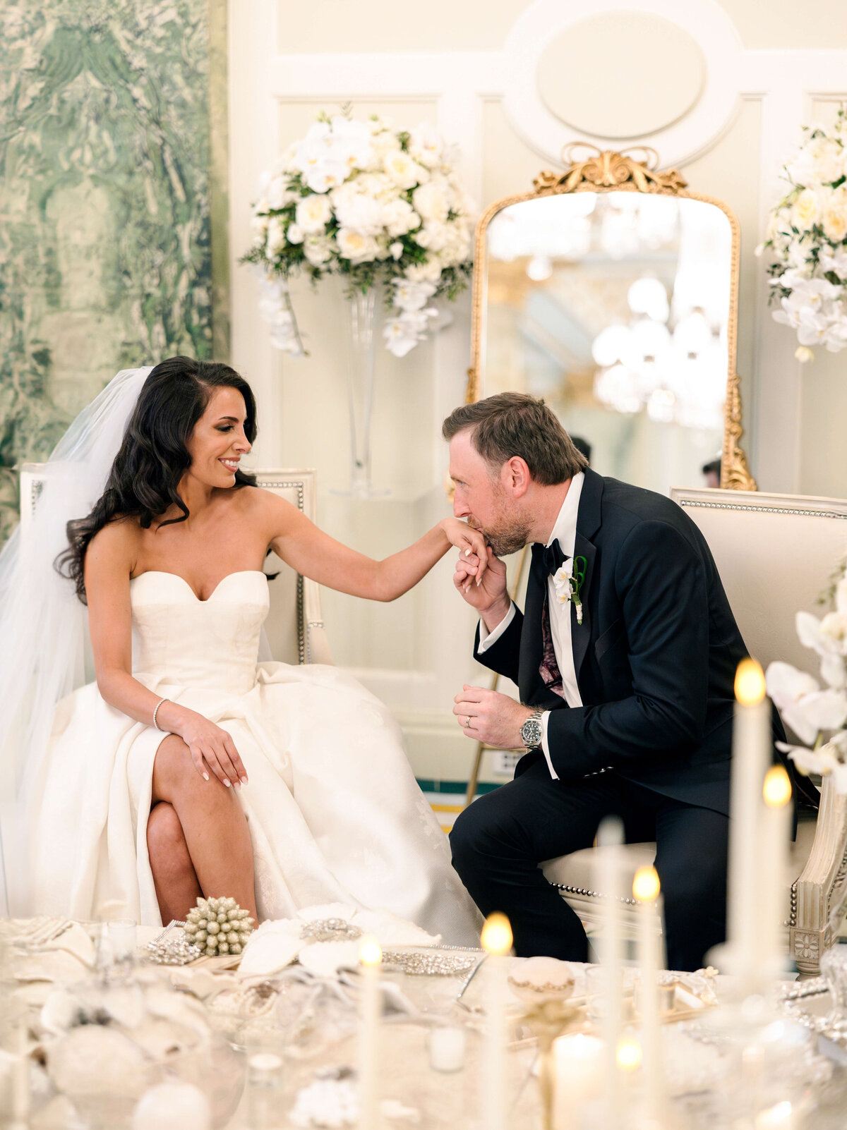 A bride and groom sit in an elegantly decorated room. The groom kisses the brides hand as they both smile. The bride wears a strapless gown and a veil, while the groom is in a black tuxedo. White flowers and candles adorn the background.