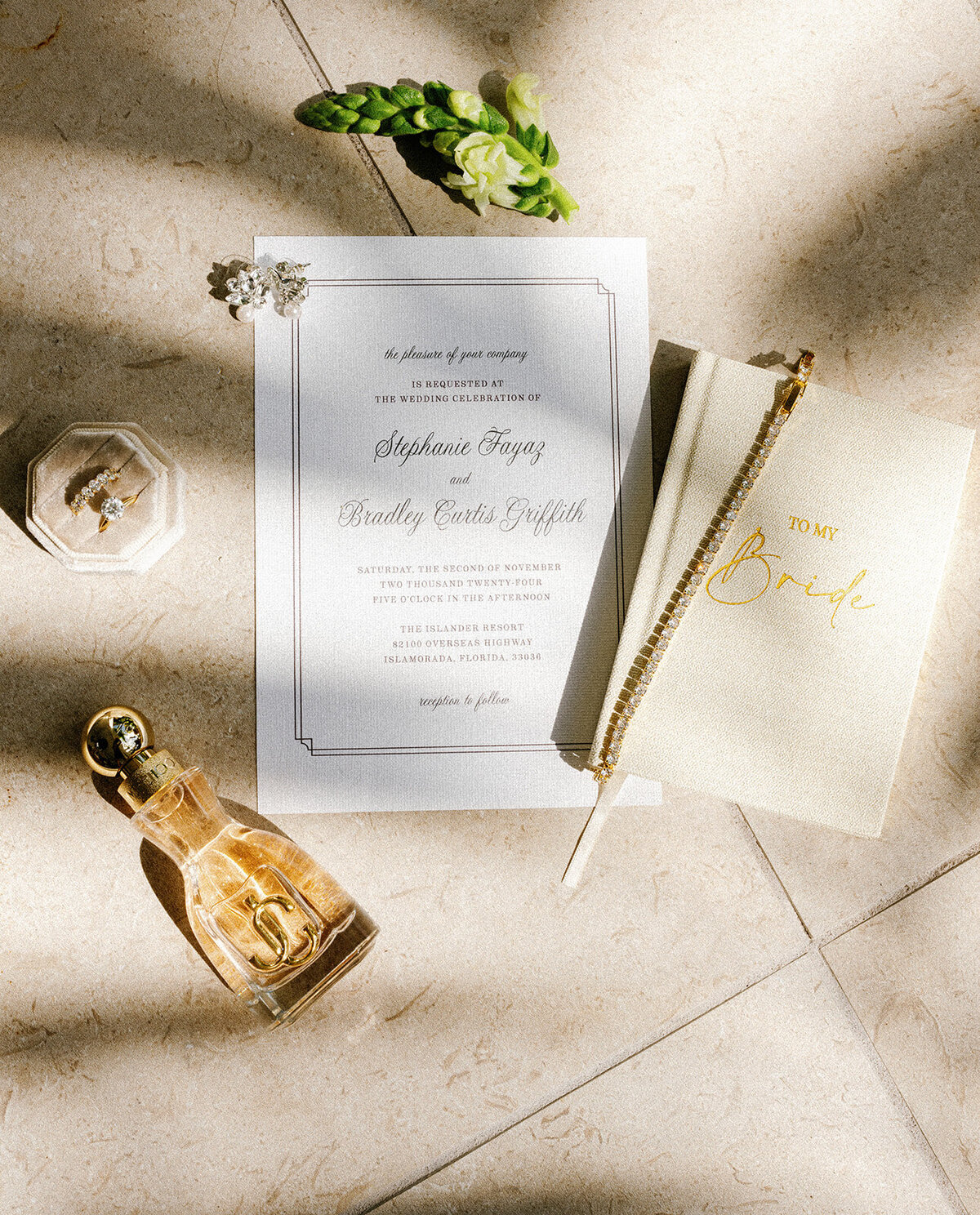 A naturally lit flat lay featuring Stephanie and Bradley’s wedding invitation, vow book, gold wedding rings, and a perfume bottle, accented by fresh white florals. Photographed by Claudia Amalia Photography in Islamorada.