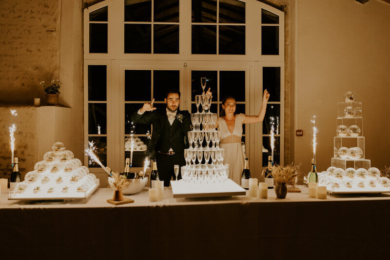 Mariés festifs derrière une structure de coupes de champagne entourée de décor de sphères rondes en verre. Prise par Laura, photographe de mariage en Vendée.