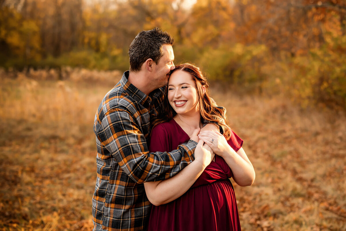 A couple embraces in an autumn field. The woman, in a maroon dress, smiles brightly, while the man, in a plaid shirt, stands behind her, gently holding her hands. Golden leaves and warm sunlight create a cozy, romantic atmosphere.