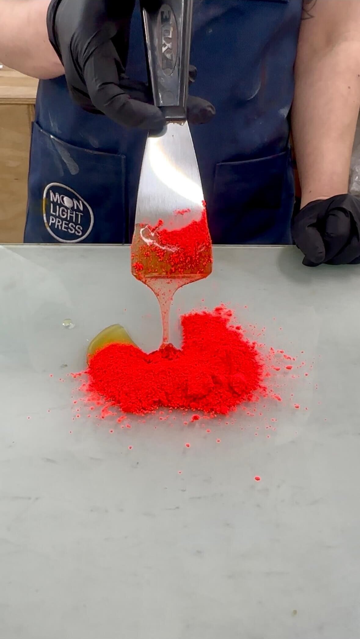 A thick stream of binder drizzles onto a pile of vibrant red pigment powder, capturing the start of hand-mixing custom ink.